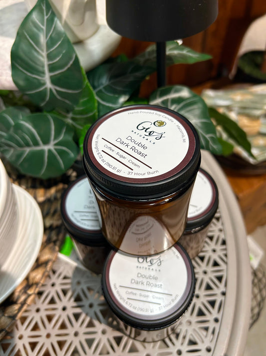 A stack of glass amber jar candles with black lids rest on a grey table with faux decorative plant material in the background. The label reads ' Gio's Naturals Double Dark Roast' and features scent notes.