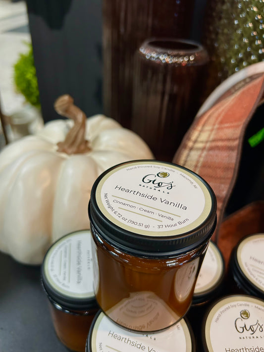A jar of Gio's Naturals andmade 'Hearthside Vanilla' candle displayed on a shelf with other jars, a white ceramic pumpkin, and decorative vases and ribbon in the background.
