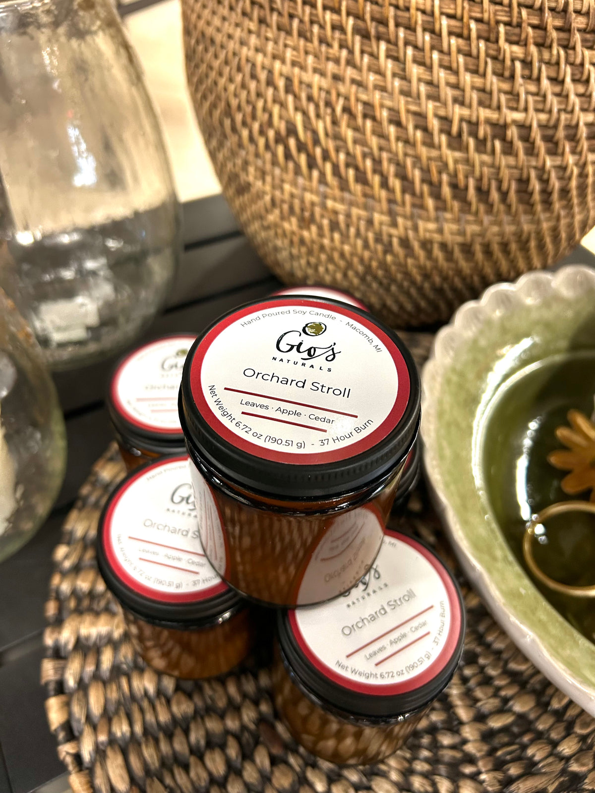 Four small amber glass jars of Gio's Naturals 'Orchard Stroll' candles arranged on a woven tray, with a green ceramic dish and a large woven basket in the background.