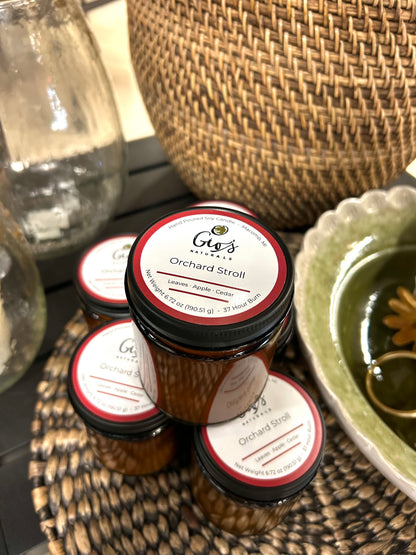 Four small amber glass jars of Gio's Naturals 'Orchard Stroll' candles arranged on a woven tray, with a green ceramic dish and a large woven basket in the background.