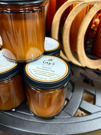 Several amber glass jars of Gio's Naturals 'Spiced Pumpkin Pie' candles with black metal lids are arranged on a dark metal tabletop. The front-center jar label reads 'Gio's Naturals Spiced Pumpkin Pie'