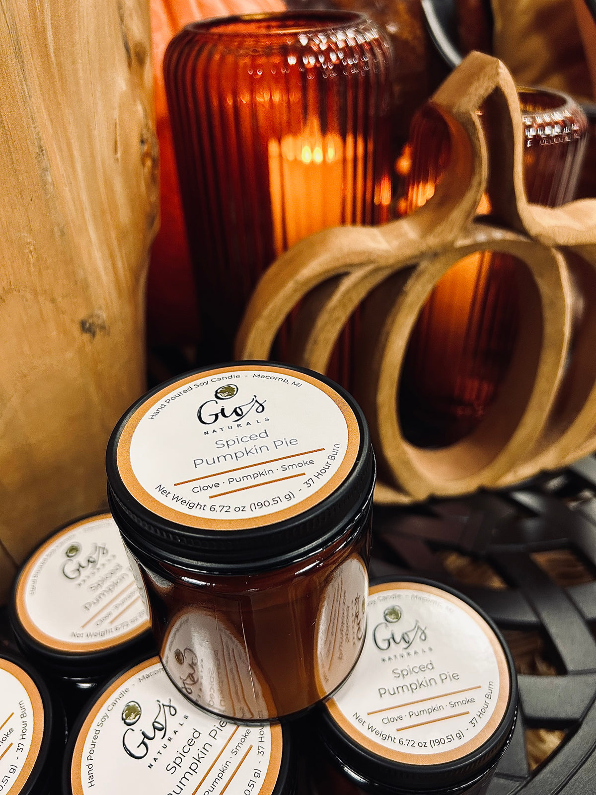 Several amber glass jars of Gio's Naturals 'Spiced Pumpkin Pie' candles are arranged on a dark metal table with rustic wooden and amber decorative props in the background.