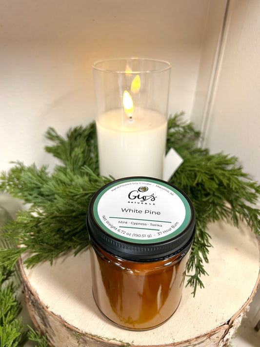 A jar candle labeled 'Gio's Natural White Pine' sits on a rustic wood slice with green pine branches, with a lit white pillar candle in the background.